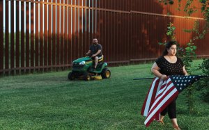 Mujer con una bandera norteamericana en su jardín y un hombre podando el pasto justo a unos pasos de la valla fronteriza.