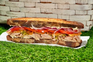 a roasted lemon and crispy chicken skin sandwich recipe on top of a paper napkin lying on fake green grass; part of a white picnic basket sits in the background