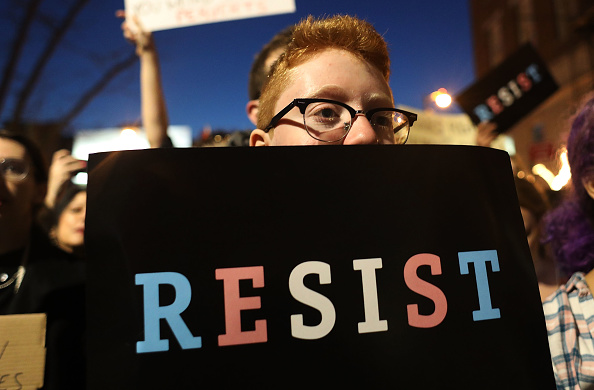 Image of a protestor with a sign that reads "RESIST" in the colors of the transgender pride flag.