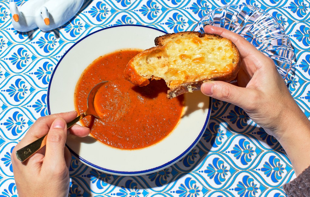 a person holding a piece of cheesy toast in one hand and dipping a spoon into tomato soup in the other