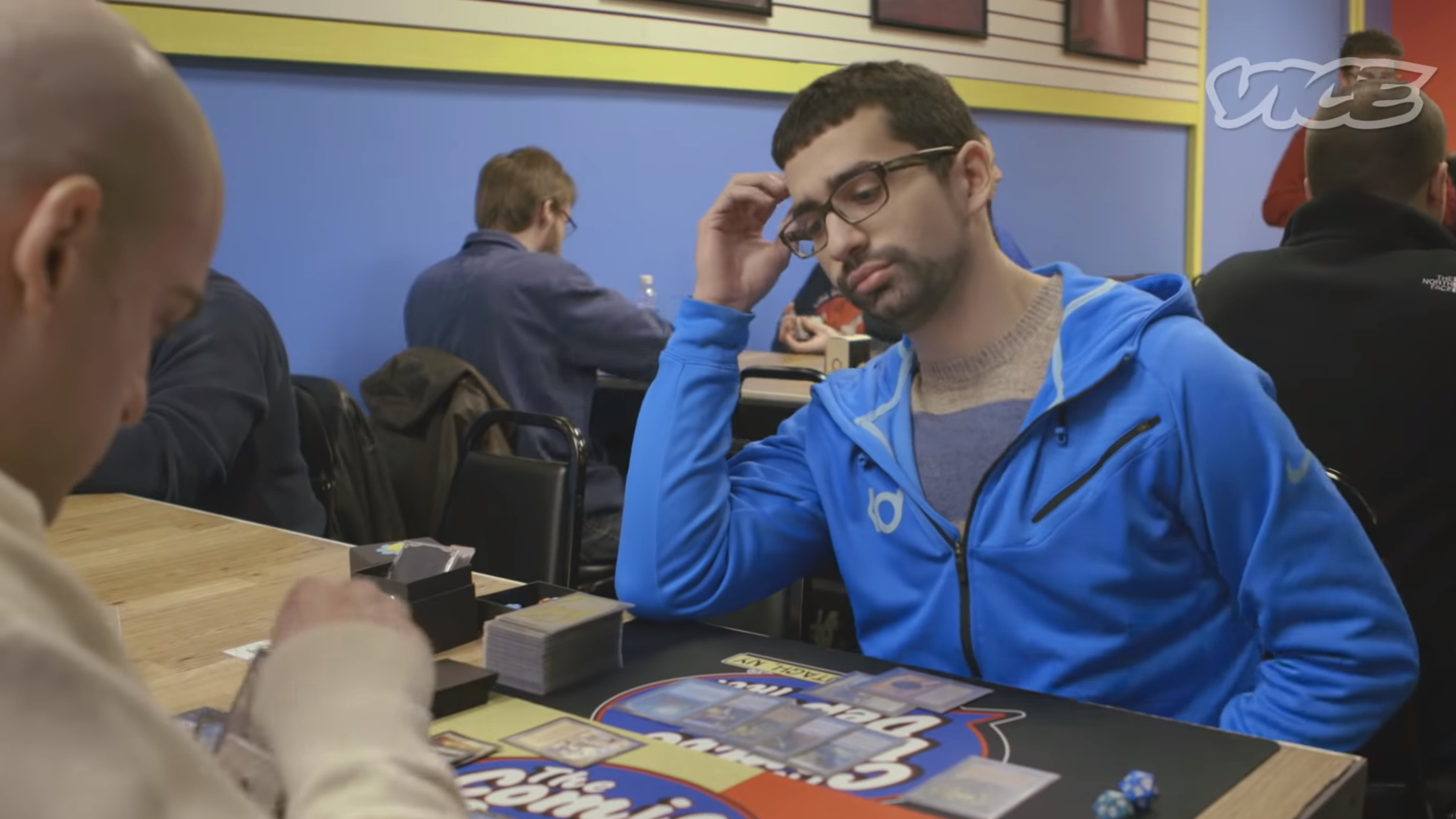 A still frame from a Vice documentary depicting a man in a blue hoodie staring contemplatively at a table covered in Magic cards.