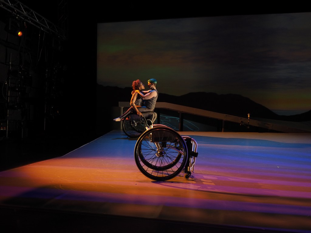 Dress rehearsal image, bathed in warm sunset hues Laurel Lawson as Venus and out of her wheelchair straddles Alice Sheppard as Andromeda in her wheelchair. They hold the intimate embrace and eye contact. An empty wheelchair is downstage.