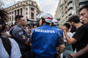 Policial com câmera na mão e outros policiais com colete azul escrito "mediador"