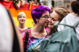 Uma mulher beijando o rosto de outra mulher com uma bandeira LGBT ao fundo