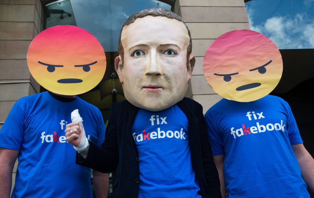 Ein Mann mit Zuckerberg-Maske zwischen zwei anderen mit wütenden Emoji-Gesichtern