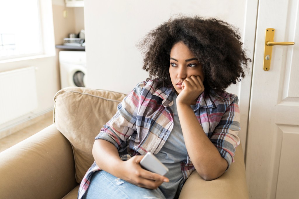 Young woman sitting on a chair with her cell phone in her hand, looking sad.