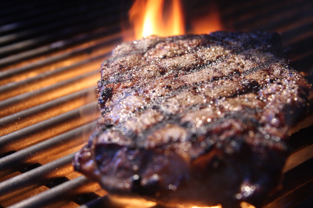 ribeye steak with crosshatched grill marks on a grill, with an orange flame in the backround