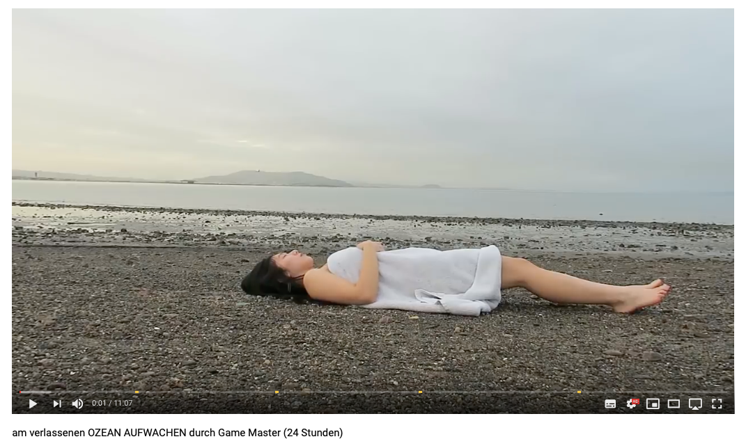 Screenshot von YouTube. Thi Lan liegt auf dem Rücken auf einem Kiesstrand. Über ihr ein Handtuch. Im Hintergrund: Meer und eine Insel