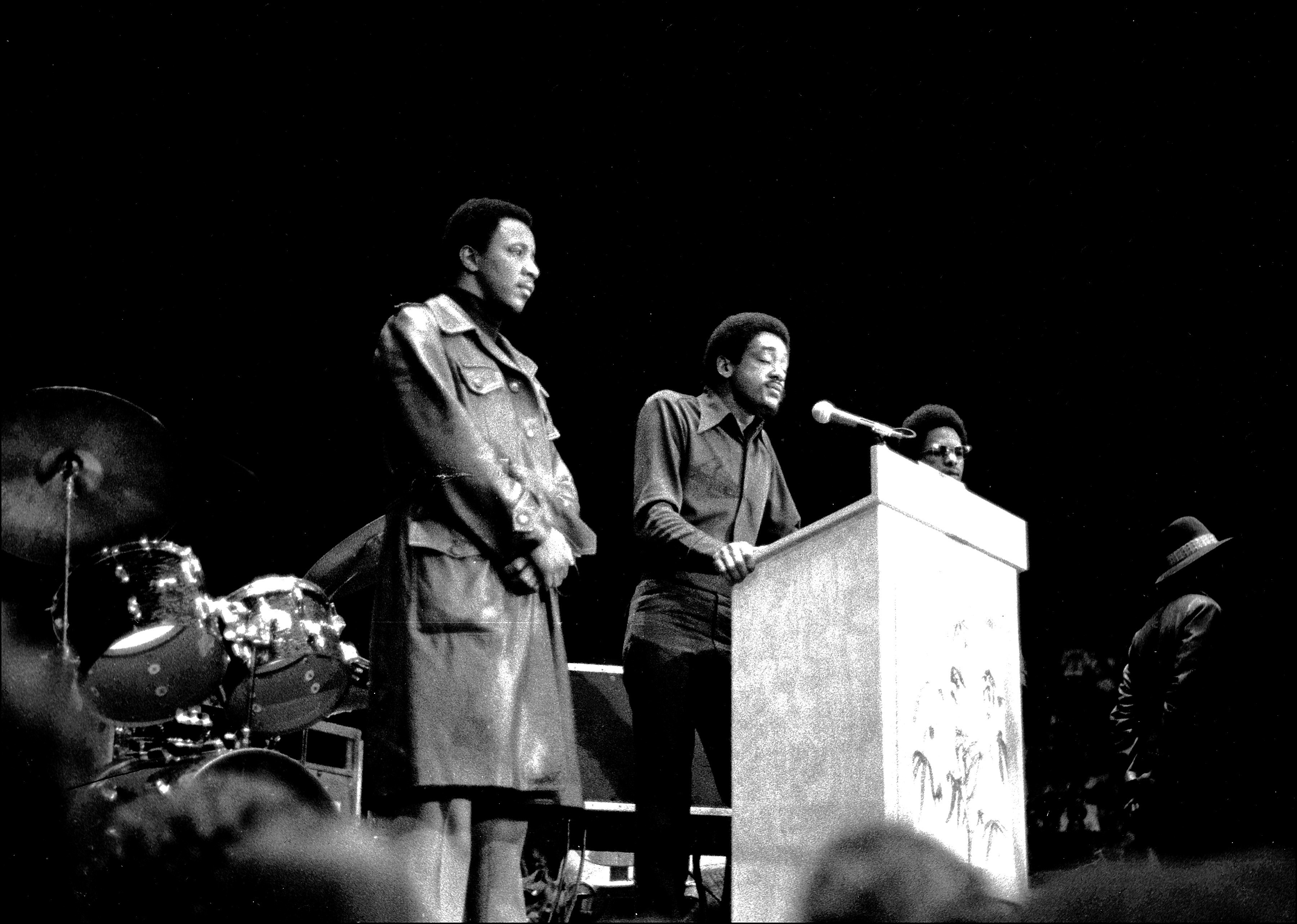 Bobby Seale speaks on a stage surrounded by bodyguards and musical equipment.