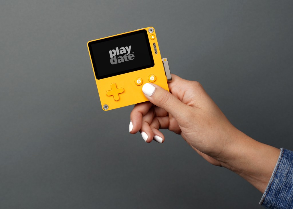 ​Image courtesy of Panic. A hand holding the Playdate handheld, a square, yellow game boy like device with a crank on the side