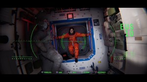 ​Image Courtesy of No Code, a woman in an orange spacesuit in a dimly lit space station, as seen through the lens of a CCTV camera. A secondary figure floats in the darkness.