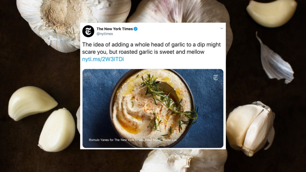 a screenshot of a tweet by the new york times for roasted garlic dip on front of a picture of heads and cloves of garlic on a black background