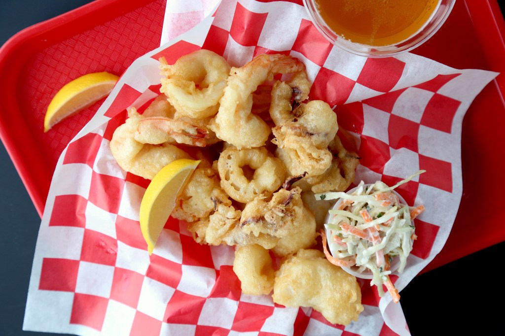 a red plastic tray holding a beer and a basket full of battered and fried shrimp and calamari, next to a cup of coleslaw, like at a seafood shack