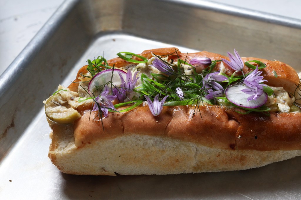 a tuna melt made in a split-top hot dog roll and topped with chive blossoms, radishes, bronze fennel, and green onion on a metal sheet tray and made by chef erik anderson of coi