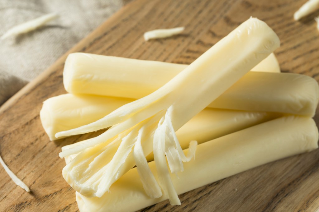 four sticks of string cheese on a wooden cutting board, one of the sticks is halfway pulled apart