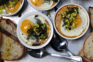 four ramekins of abra berens's eggplant and tomato stew with baked, coddled eggs on top of a metal baking sheet, surrounded with spoons and slices of toast