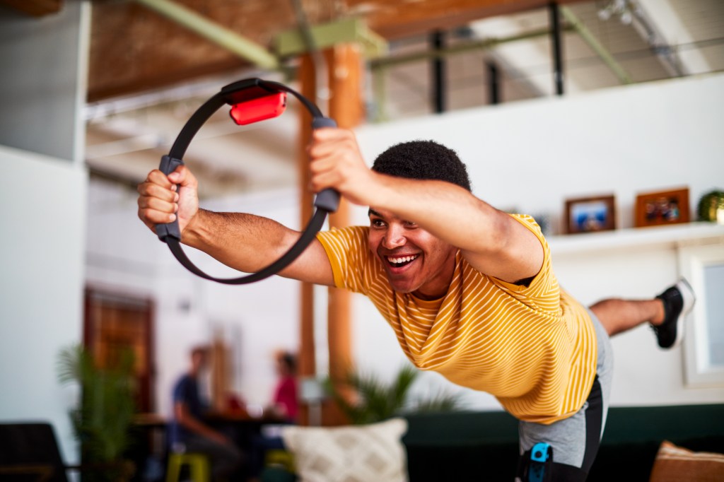 A young man with dark skin and black hair in a yoga pose, holding a black ring with a JoyCon attached to it in front of him as he stretches his right leg behind him.