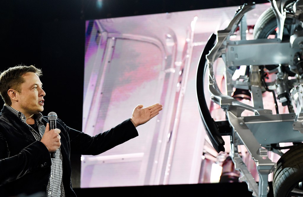 Elon Musk waving his hands at a car made by Tesla, his car company.