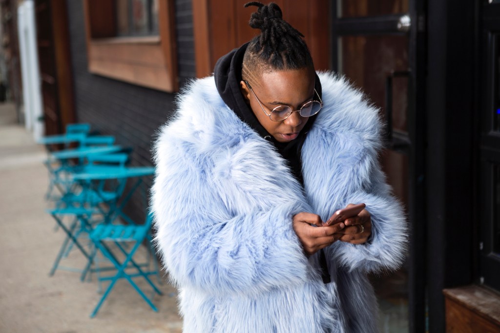 A transmasculine person with a furry blue coat checking his phone on the sidewalk