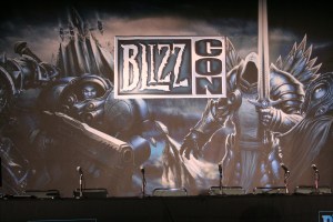 Photo of an BlizzCon panel, mics on a table with a black tablecloth, with a "BlizzCon" backdrop featuring characters from Starcraft and Diablo.