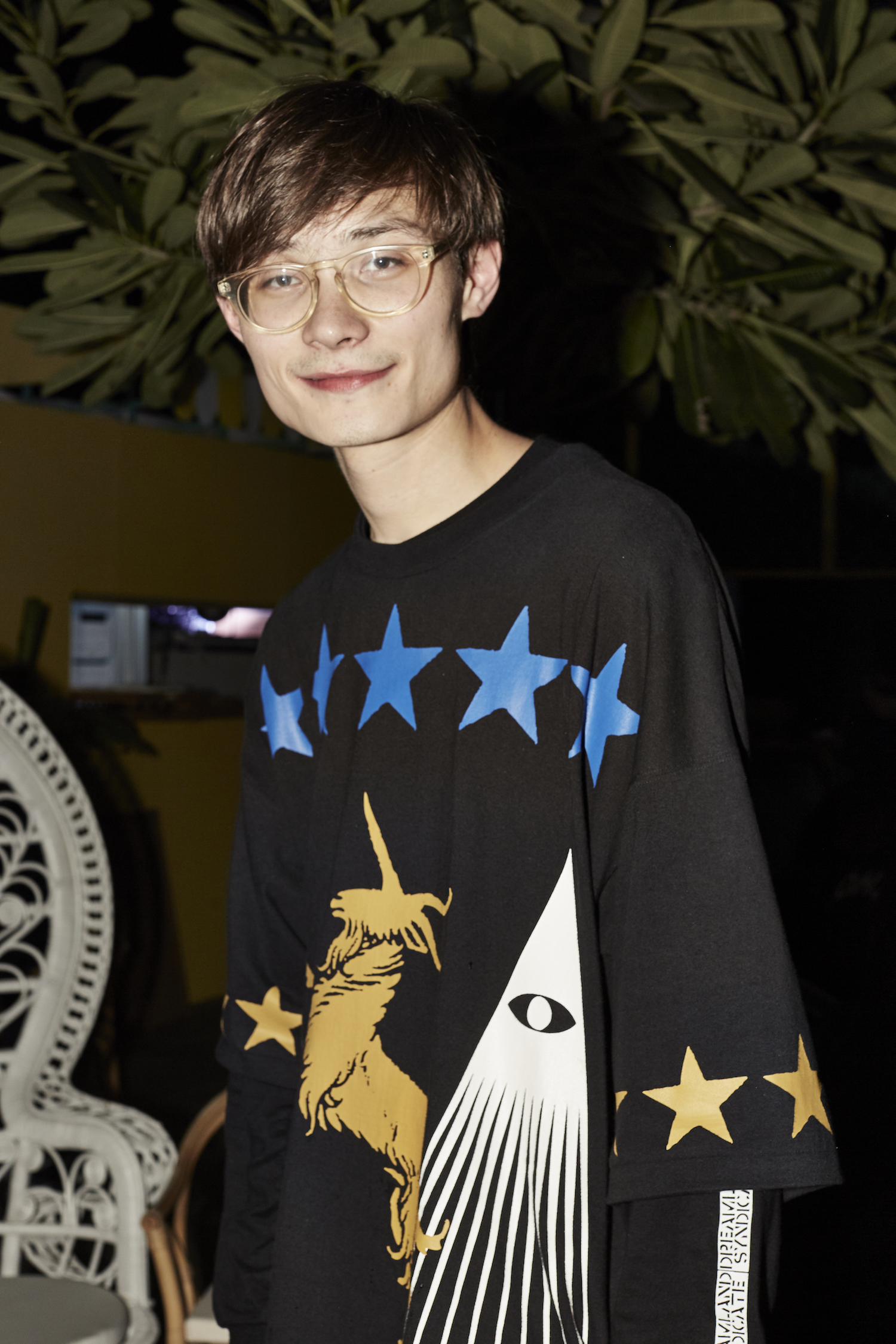 A young man wearing glasses smiles and looks towards the lens.