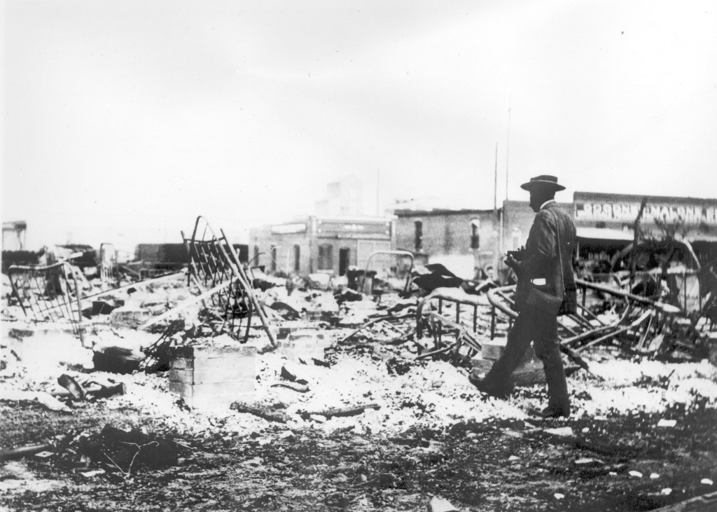 Photograph of an African-American man with a camera looking at the skeletons of iron beds which rise above the ashes of a burned-out block after the Tulsa Race Riot, Tulsa, Oklahoma, 1921. (Photo by Oklahoma Historical Society/Getty Images)