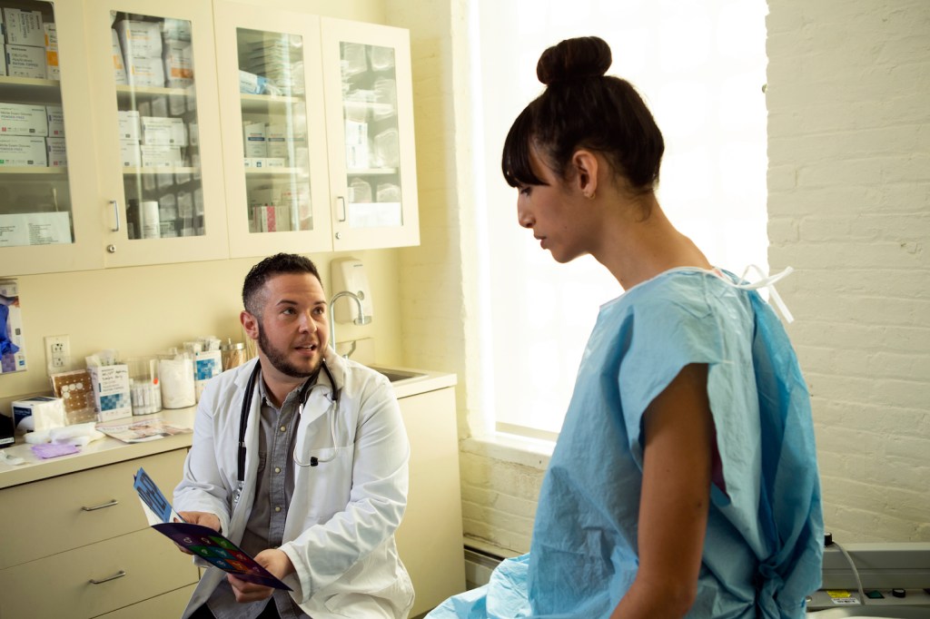 A transgender woman in a hospital gown speaking to her doctor, a transgender man, in an exam room