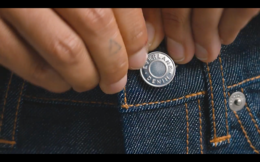 An closeup image of a person button their jeans; the word "Everlane" is visible on the button.