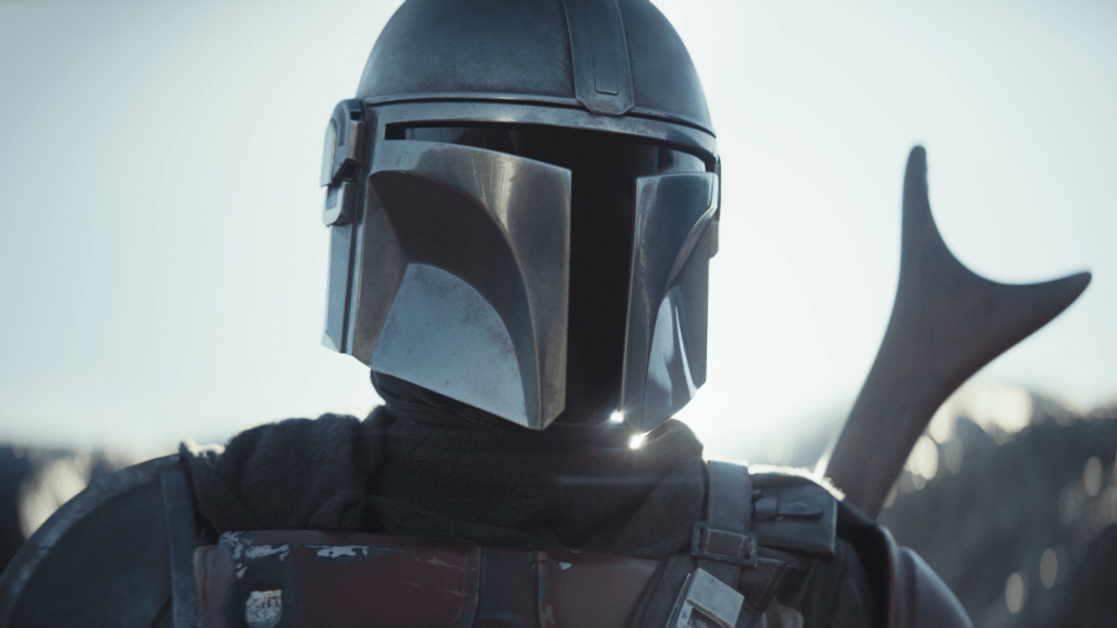 Screenshot from The Mandalorian. Close up of the Mandalorian's bust framed against a bright yet overcast sky, wearing the iconic mandalorian helmet with a "T" shaped visor.