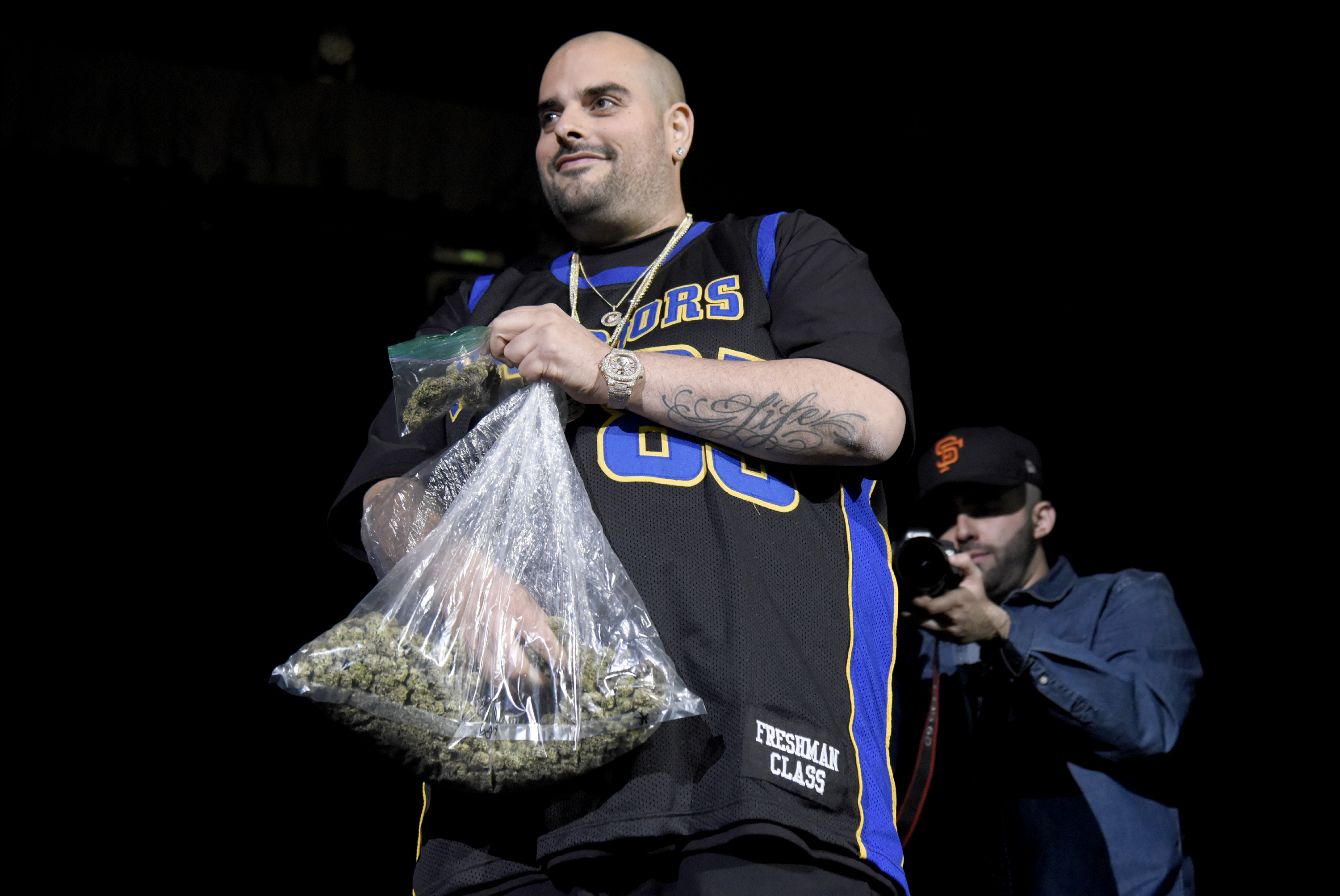 Berner stands onstage at a concert holding a big bag of weed.