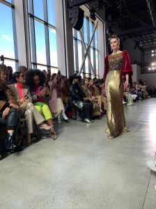 A model from the Badgley Mischka FW2020 runway show wearing a dress with red velvet sleeves and an embroidered gold body.