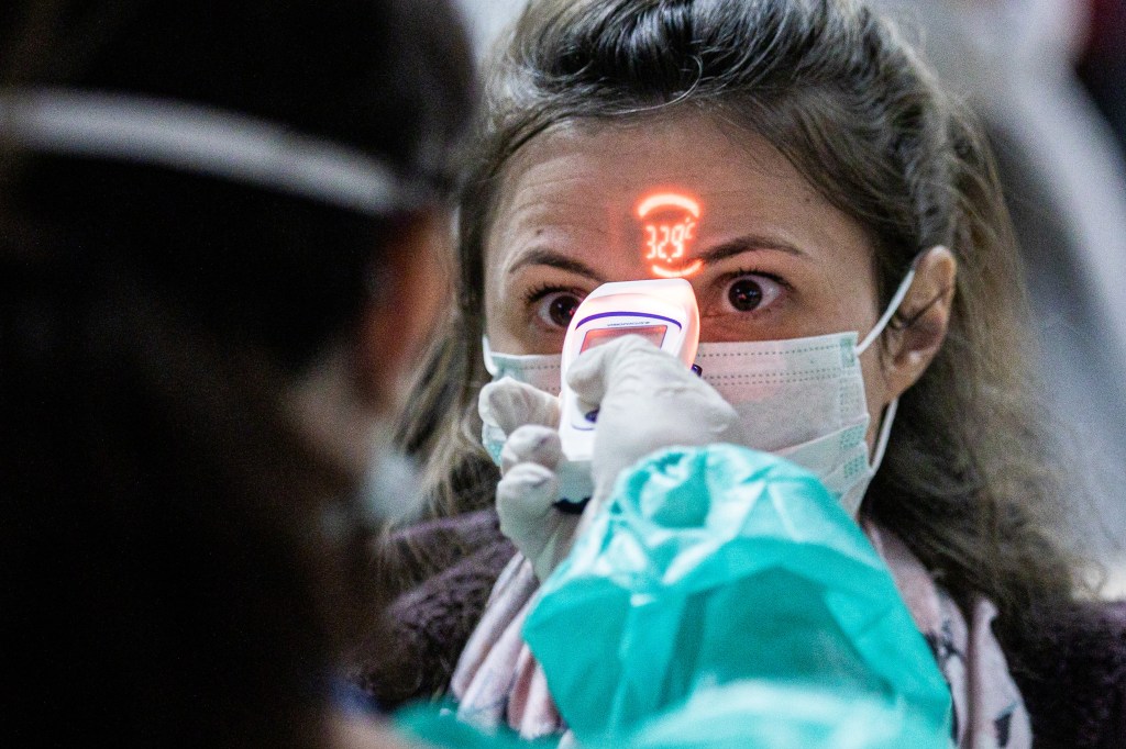 A woman in a paper face mask has her forehead scanned with a digital thermometer by a health worker; her temperature is shown displayed on her forehead.