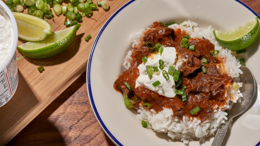 a bowl full of freezer-friendly, bean-free red chili over white rice with lime and sour cream