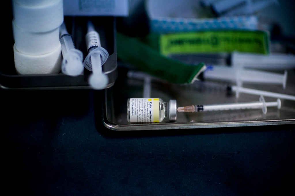 A vial of ketamine is shown sideways on a table with a syringe in it.