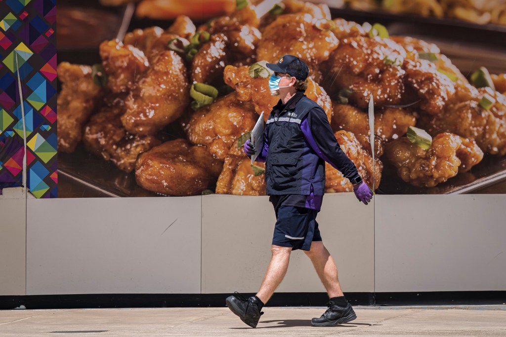 A person in a FedEx uniform wearing gloves and surgical mask walks past an advertisement for chicken.