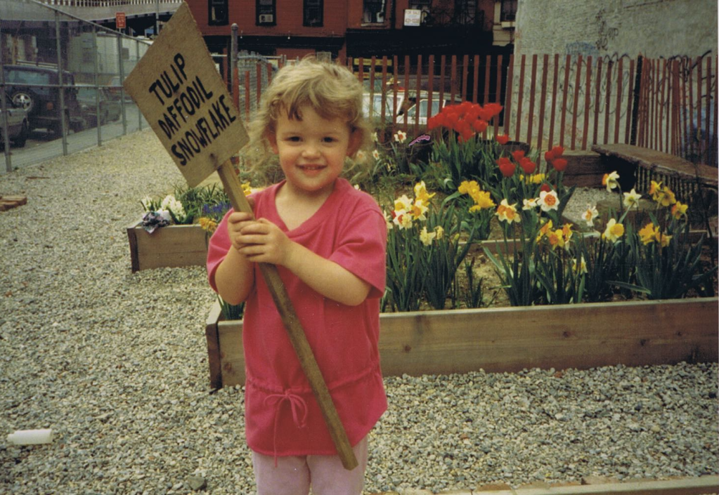 The author shown in the community garden bearing a sign for flowers amidst the flowers themselves