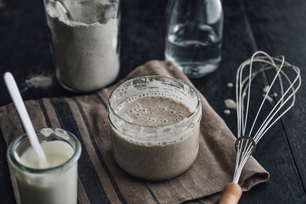 A jar of sourdough starter next to a whisk and jar of flour