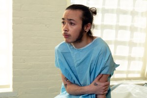 A genderqueer person sitting in a hospital gown sitting in an exam room and looking worried