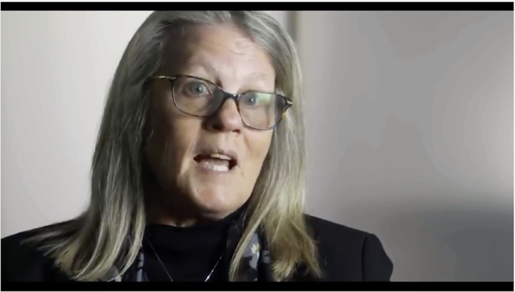 Judy Mikovits speaks in front of a grey background.