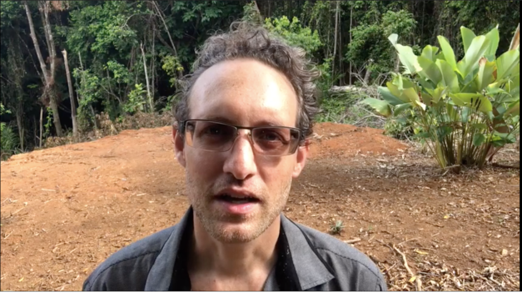 Zach Vorhies speaks in front of some green plants about his working promoting Judy Mikovits