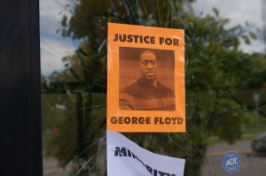 an orange sign with a picture of george floyd that says "justice for george floyd" on a cracked glass door