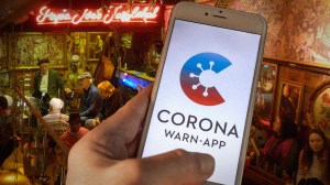 ​Menschen sitzen in einer Bar, davor hält jemand ein Smartphone mit der Corona-Warn-App. Die App wurde von Datenschützern kritisiert, gilt aber jetzt als sicher.