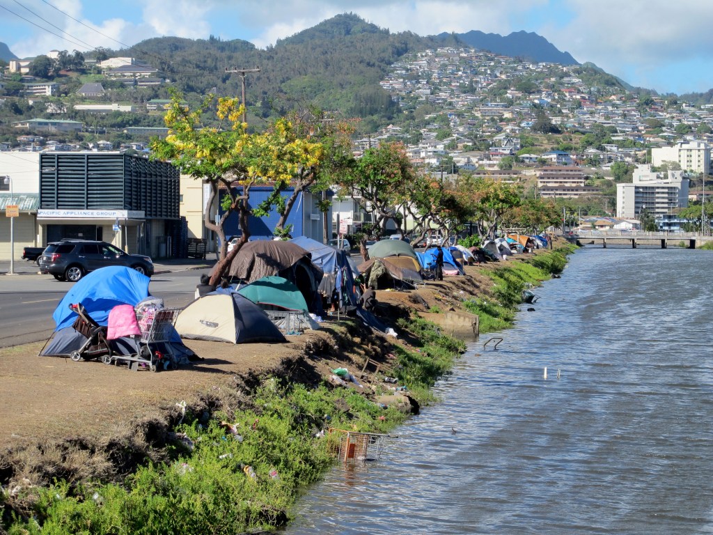 In this Thursday, June 11, 2015 photo, homeless people and their tents line a canal in Honolulu. Hours after a city crew cleared the banks of the canal, the homeless people that had been living there moved right back to the riverside, leaving some wonderi