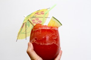 a hand holding a glass of frozen watermelon slushie cocktail with a neon green straw, yellow cocktail umbrella, and a slice of fruit