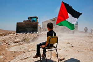 Child sitting on a chair with a Palestinian flag in a deserted area, with a house and a bulldozer on the background.