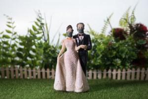 A cake topper of a bride and groom in facemasks is shown resting on fake grass, against a background of a fake picket fence.