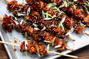 wooden skewers of homemade sesame chicken, covered in a ketchup and soy glaze and finished with sesame seeds and scallions, on top of a metal baking tray