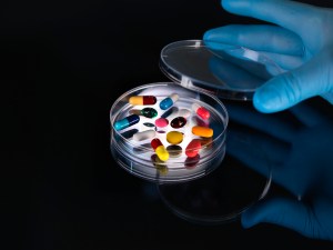 A blue-gloved hand opens the lid of a petri dish containing colorful pills with a dark background