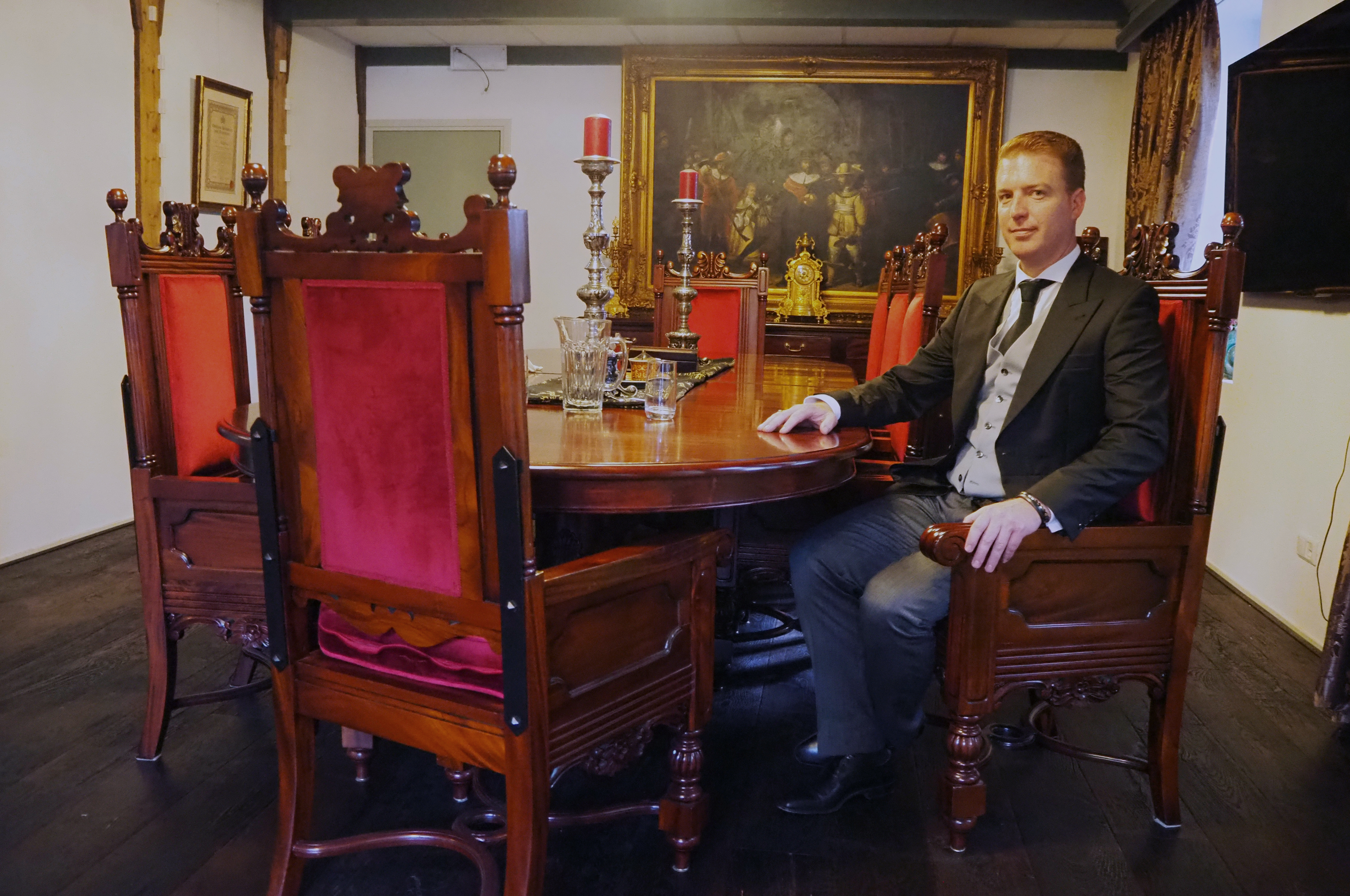 André Meester sitting at a large antique table surrounded by paintings.