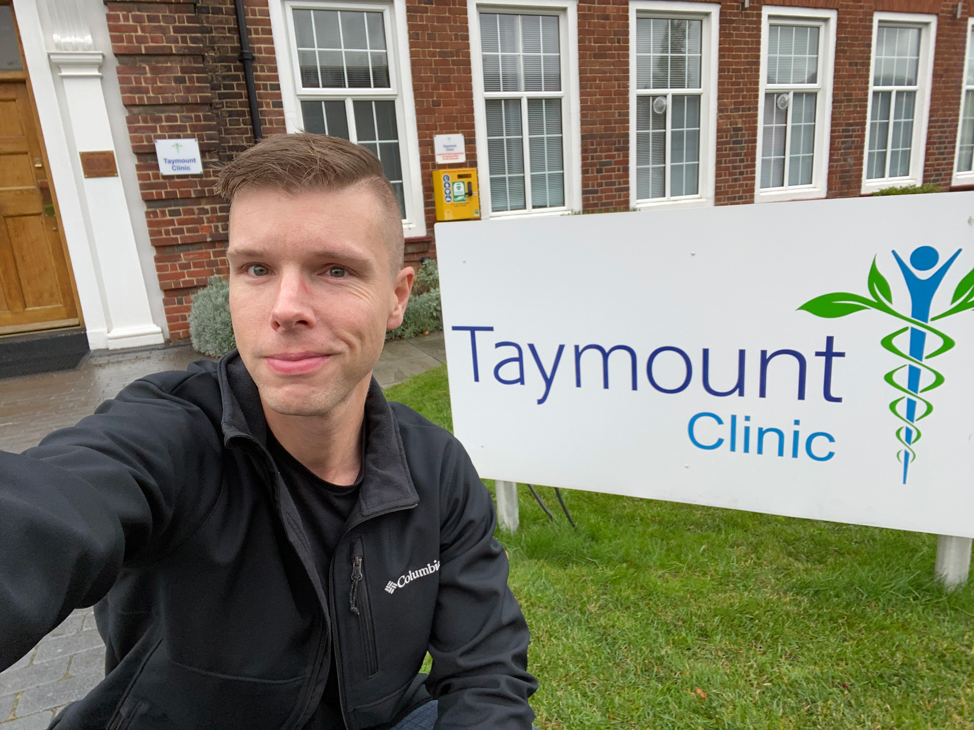 Josiah Pugh. Man with blond short hair and blue eyes, taking a selfie in front of the Taymount clinic sign.